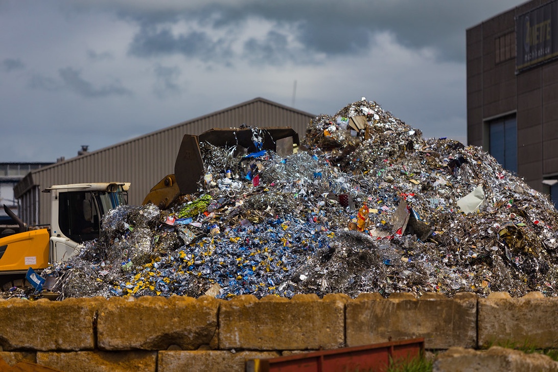 En stor hög av strimlat metallskrot och återvinningsbart material vid en industriell återvinningsanläggning. Högen består främst av metallavfall inklusive aluminium och andra metaller i olika färger. Industribyggnader och utrustning syns i bakgrunden under en mulen himmel, vilket indikerar en kommersiell återvinningsverksamhet.